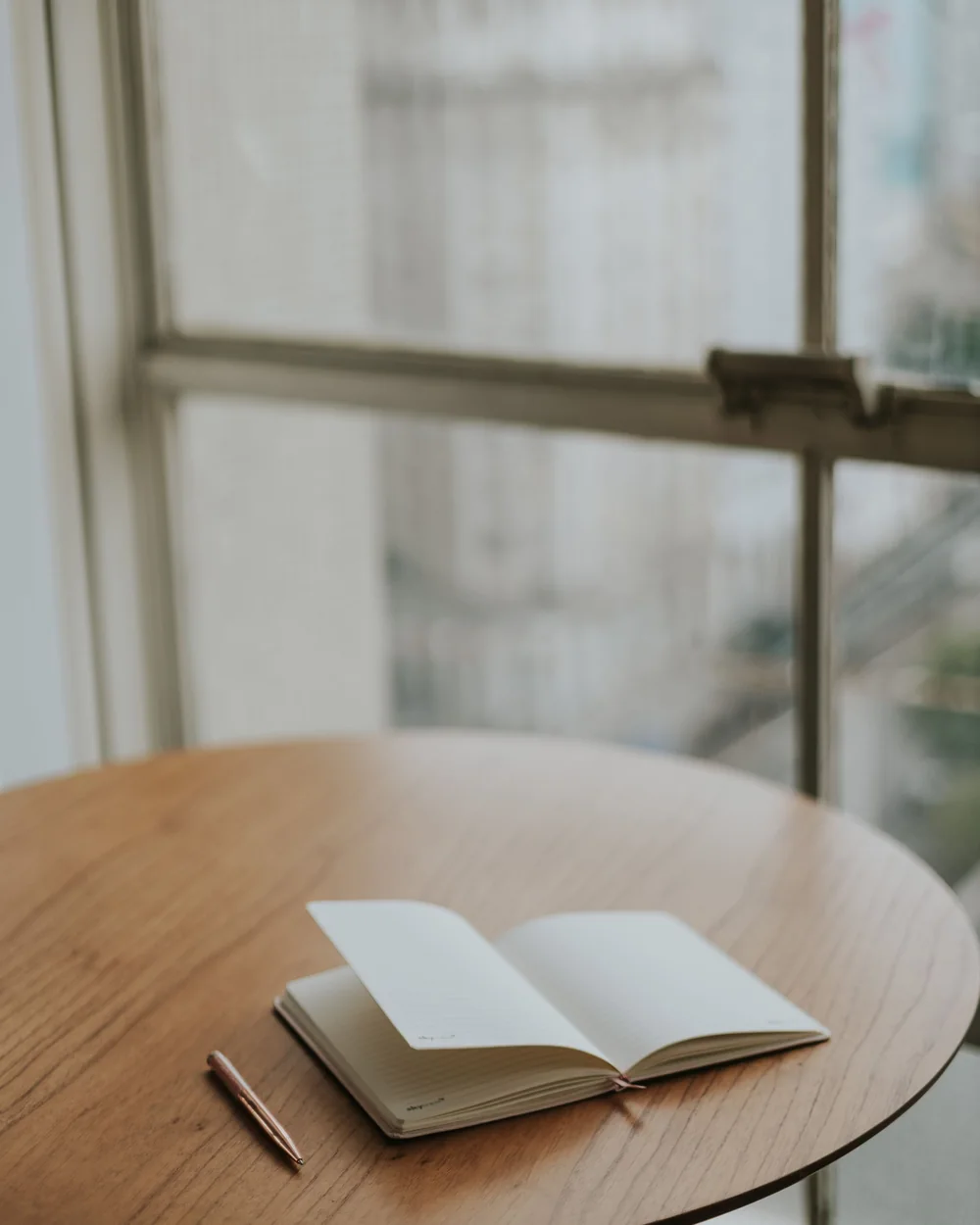 Un carnet ouvert et un crayon posés sur une table ronde, près d'une fenêtre; image d'ambiance